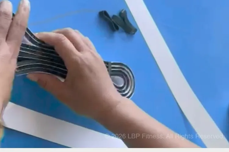A realistic documentary-style photograph from a factory floor, showing a close-up of a worker's hands inserting a sheet of protective wax paper between the layers of a rolling red latex resistance band